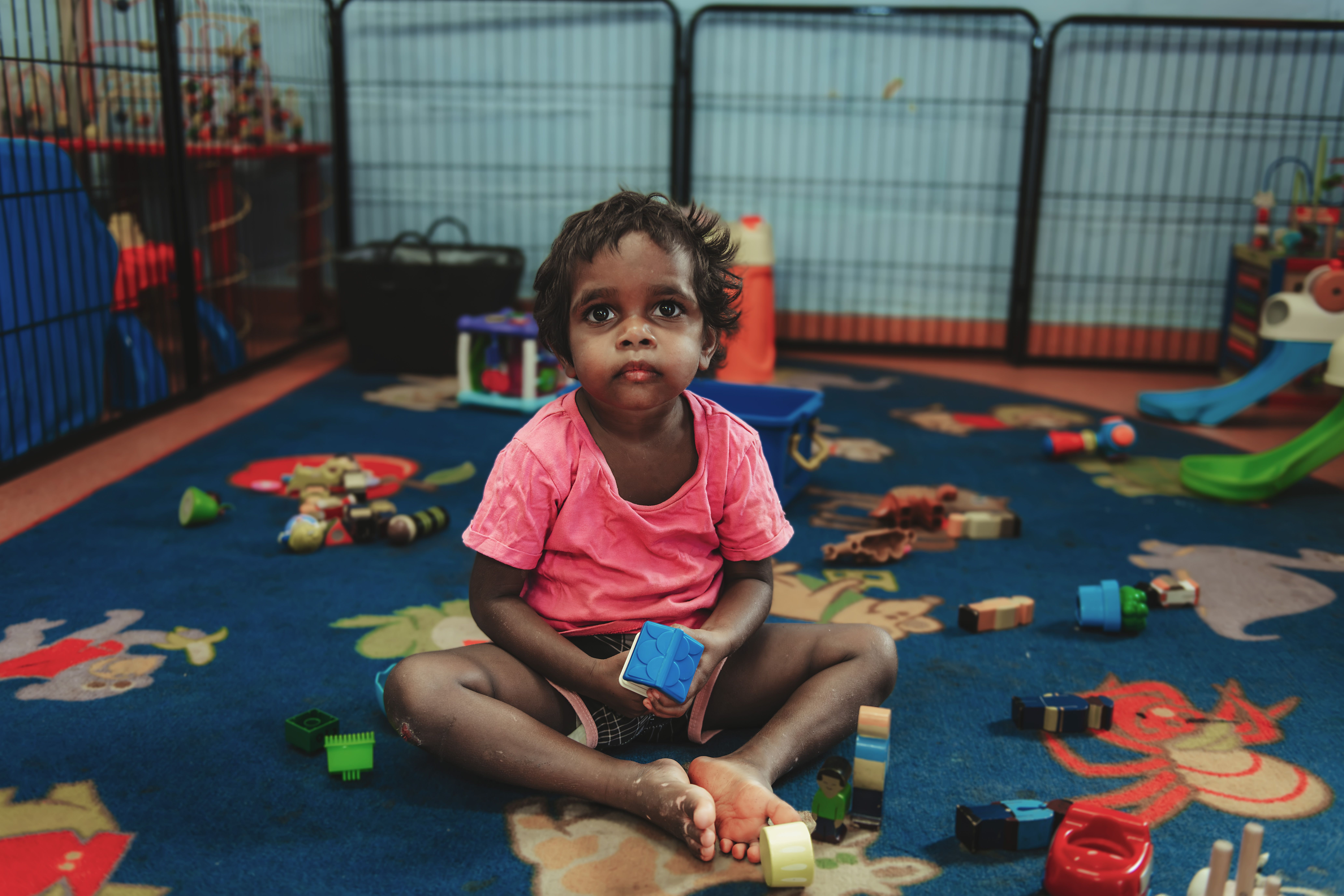 Young child sitting on a rug surrounded by toys, holding a blue block