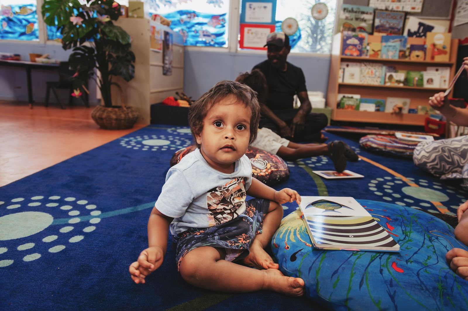 Young boy sitting on a rug, looking at the camera with a book in front of him