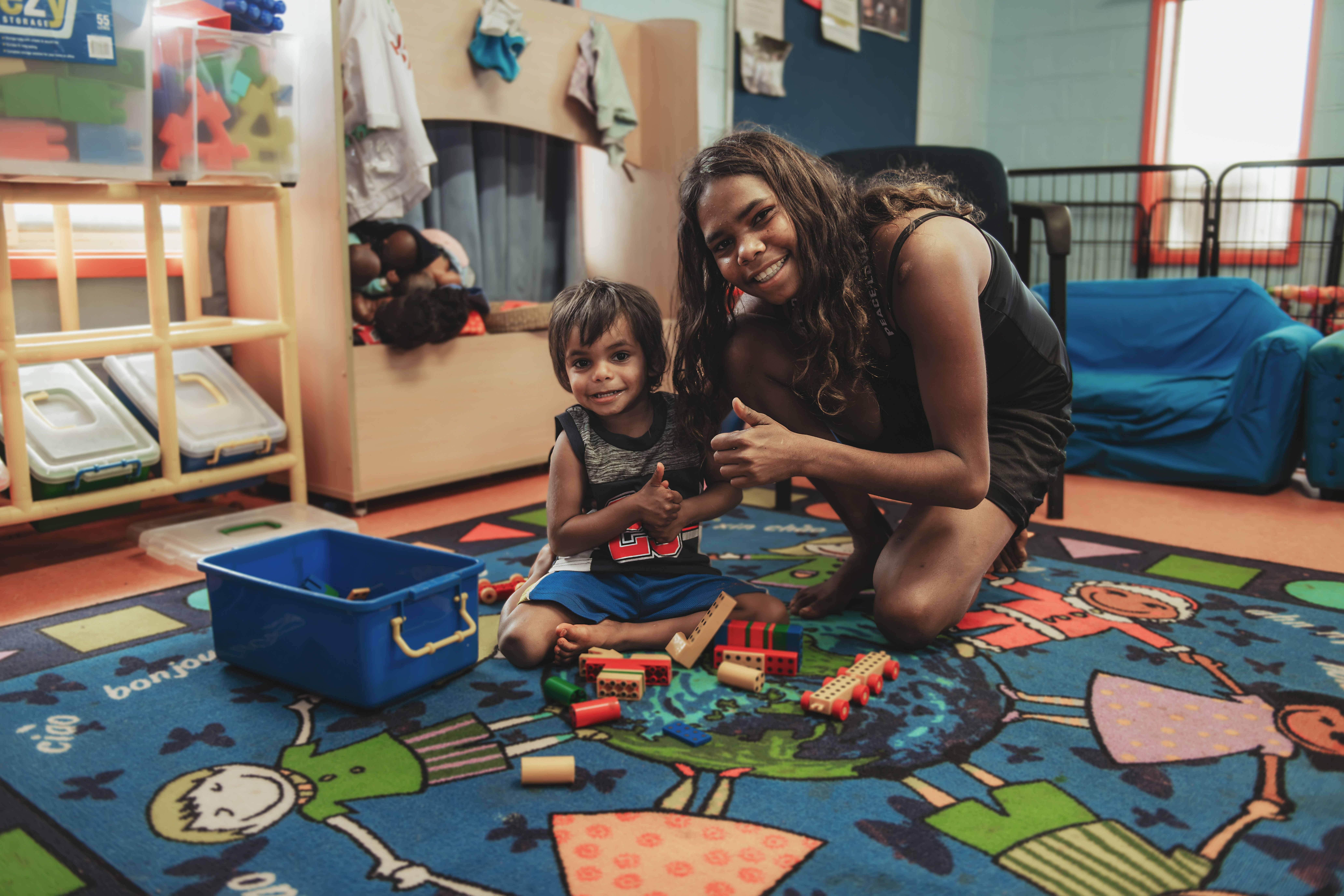 Photo of girl and younger boy playing with toys, smiling at the camera with their thumbs up