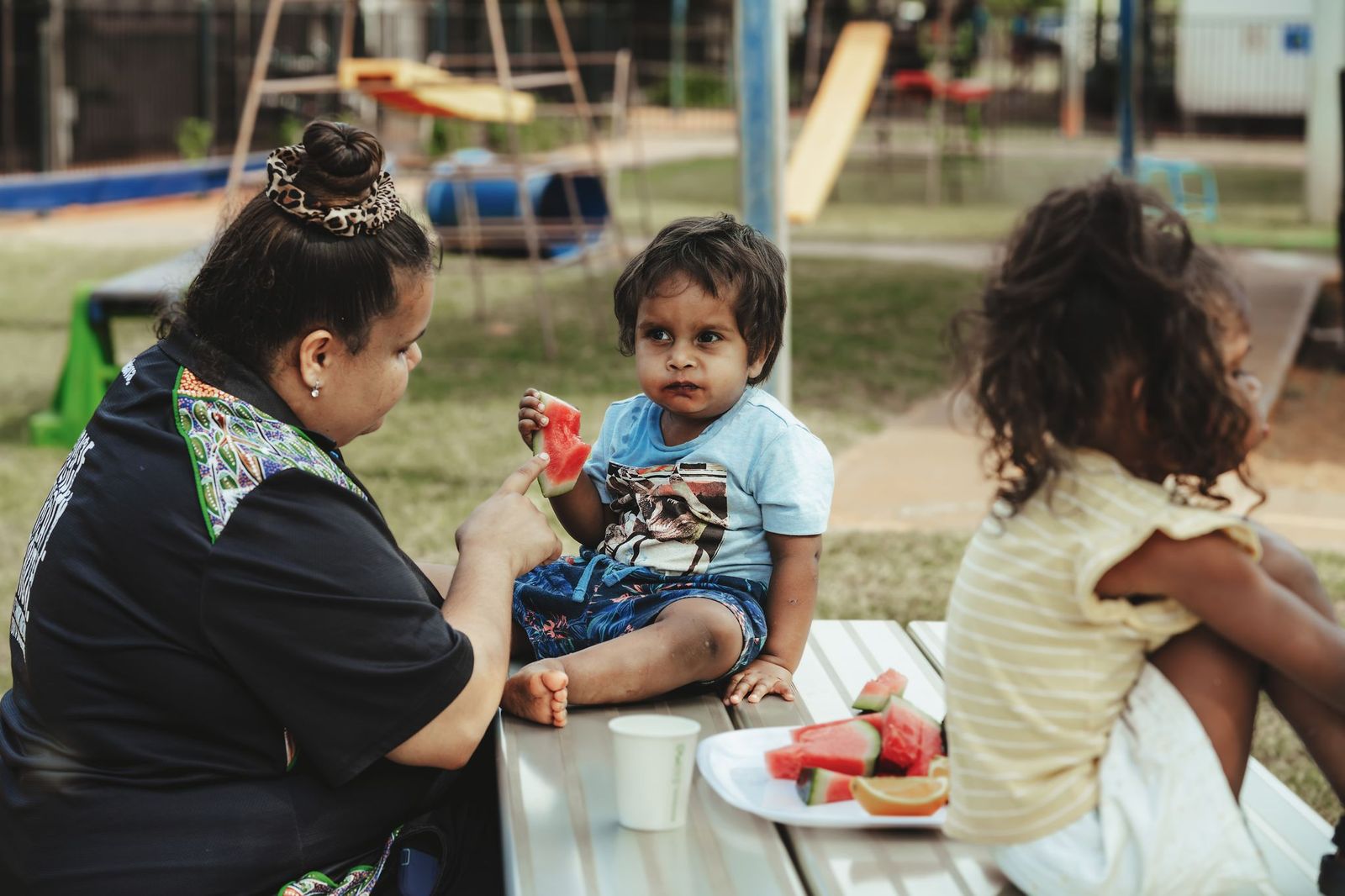 Young boy with a carer sitting on a table eating watermelon