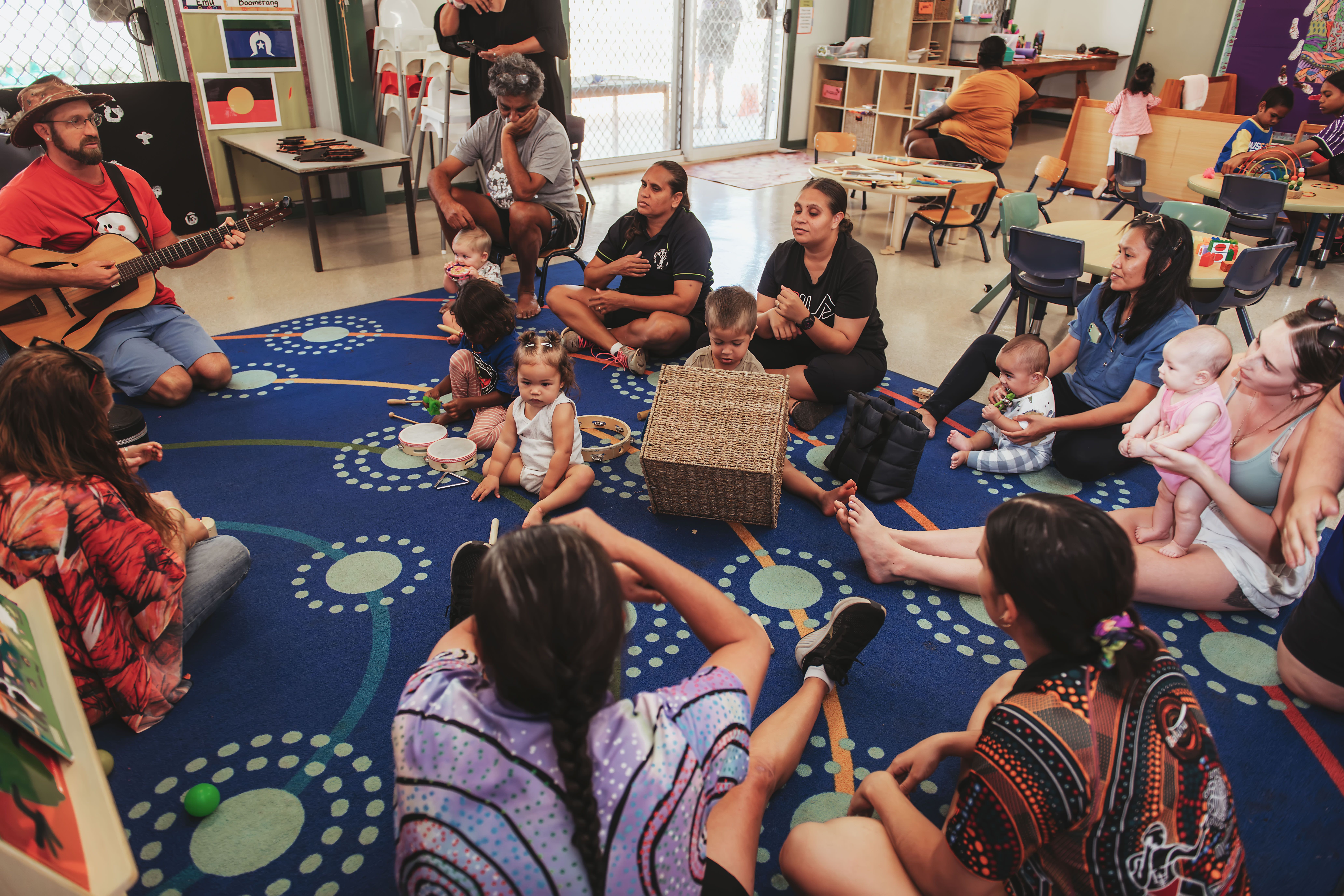 Young children, educators, parents and carers sitting around a circle listening to guitarist